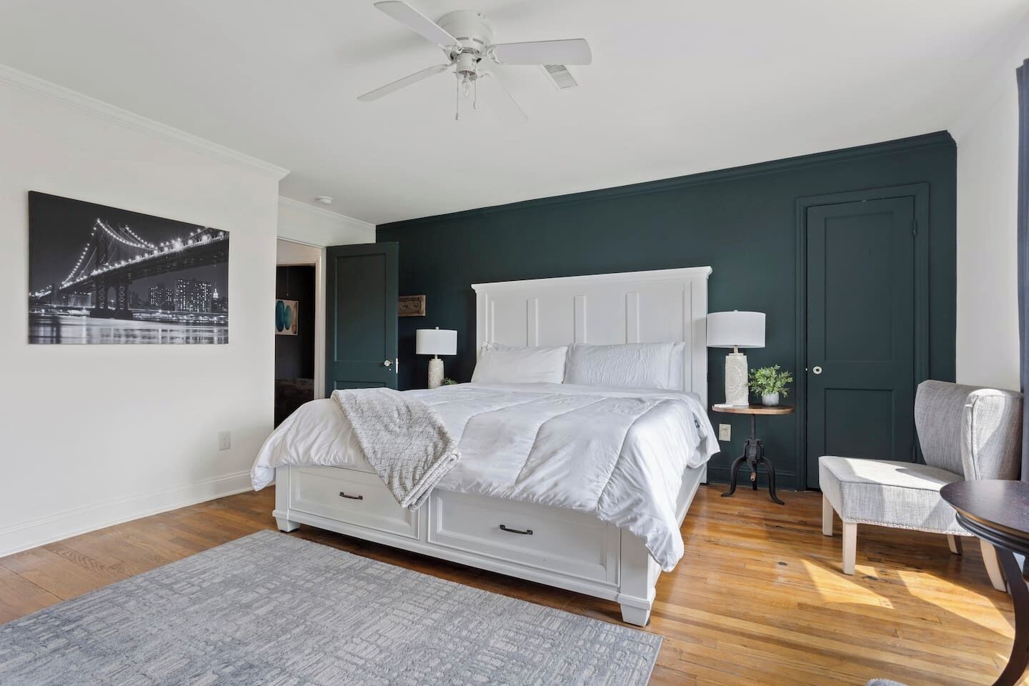 A bedroom with a white bed, dark walls, a ceiling fan, a night view of a bridge, and a small table with a lamp.