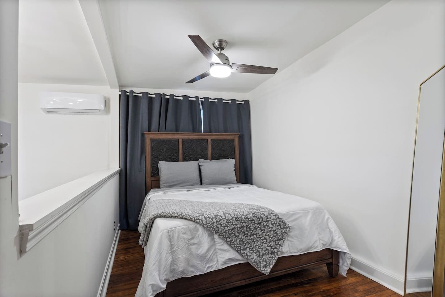Una habitación pequeña y estrecha con una cama individual, un ventilador de techo y una ventana con cortinas.