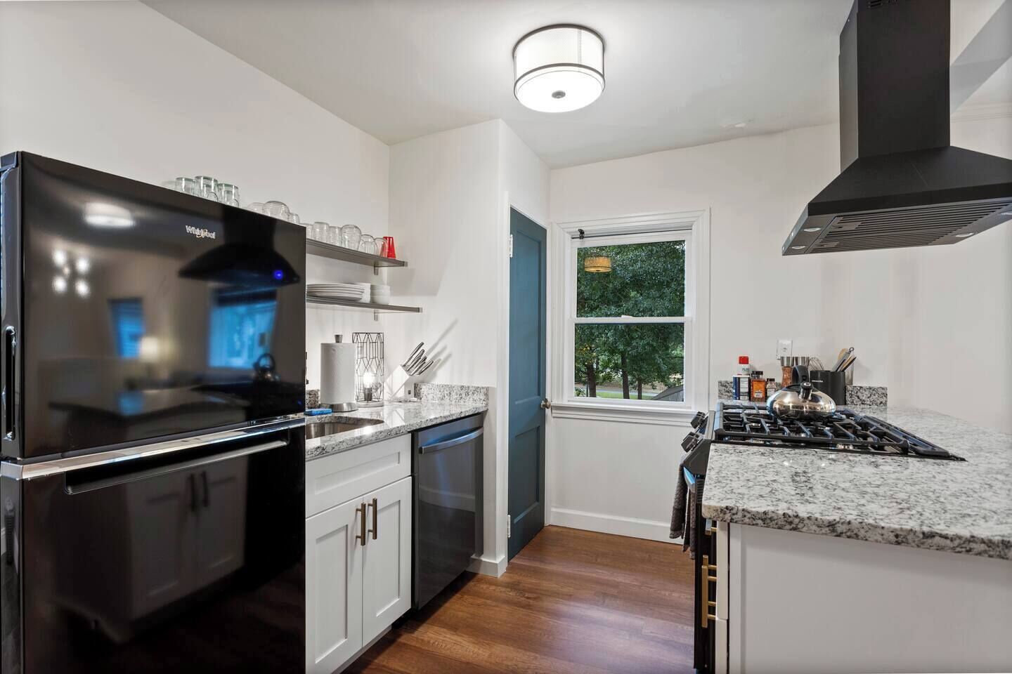 Una cocina moderna con un refrigerador negro, electrodomésticos de acero inoxidable, gabinetes blancos y una ventana con vista a árboles.