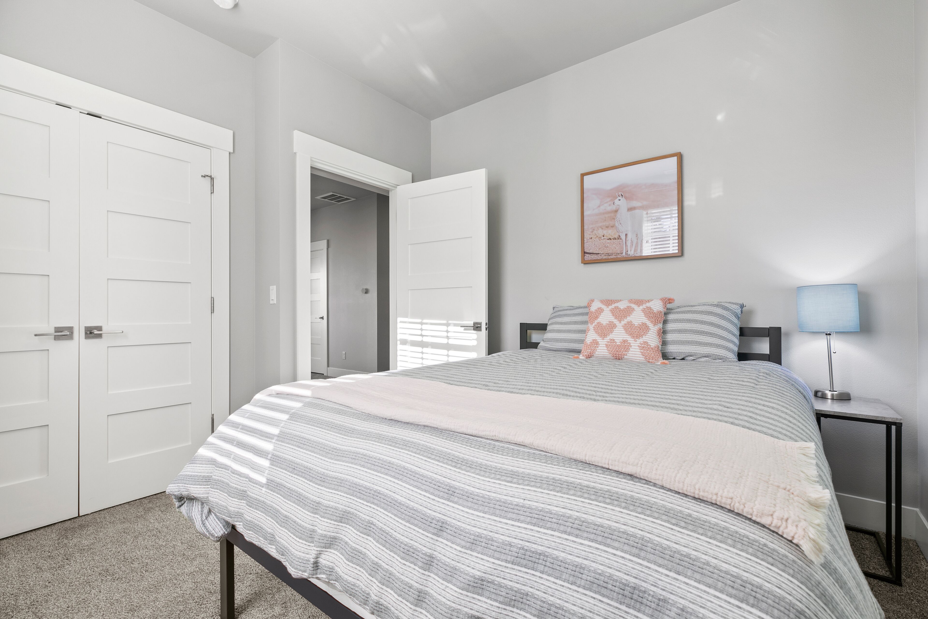 A bedroom with a bed, a nightstand, a lamp, a framed picture on the wall, and a door leading to another room.