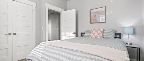 A bedroom with a bed, a nightstand, a lamp, a framed picture on the wall, and a door leading to another room.