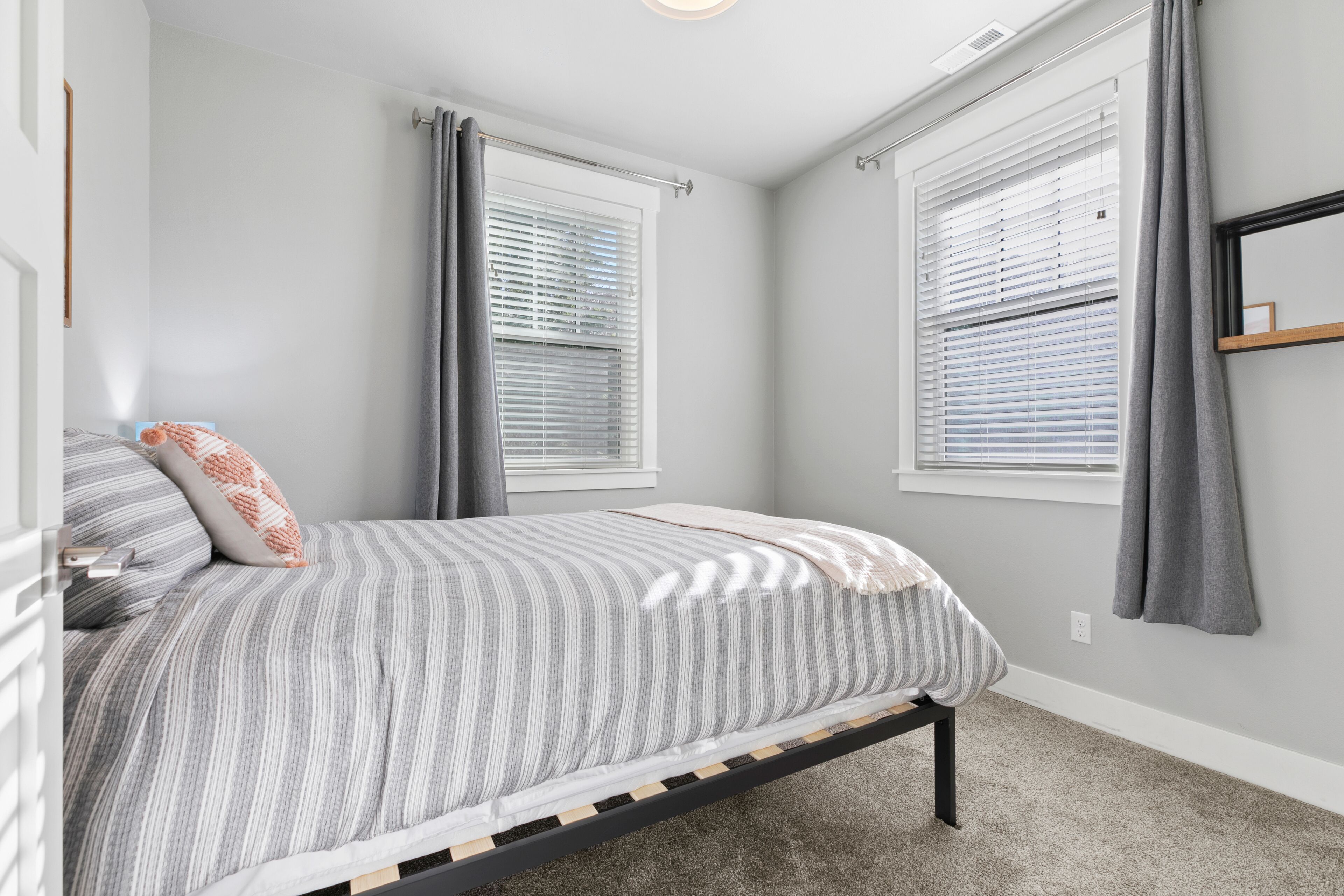A bedroom with a bed, two windows with blinds, a bedside table, and a carpeted floor.