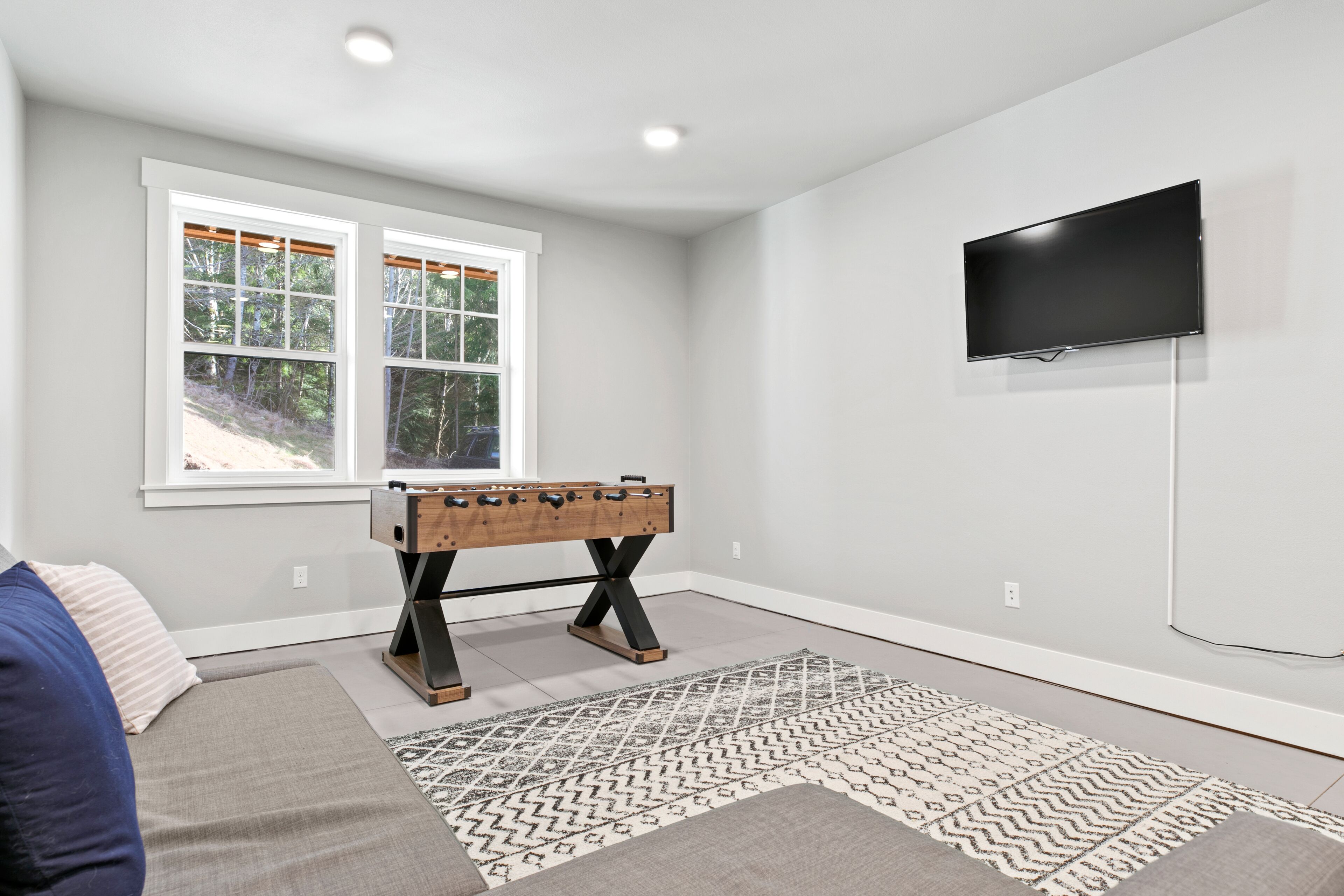 A room with a foosball table, a TV mounted on the wall, and a window with a view of trees.