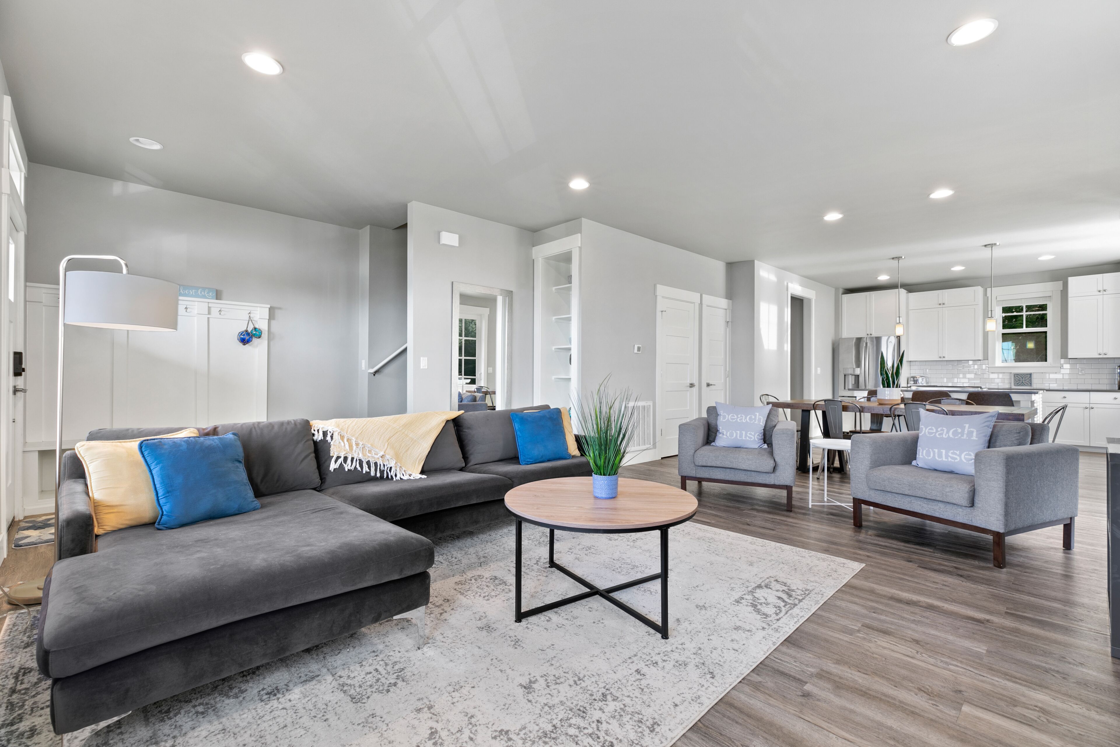 A modern living room with a gray sectional sofa, a round coffee table, and two armchairs. A kitchen is visible in the background with white cabinets and a dining area.