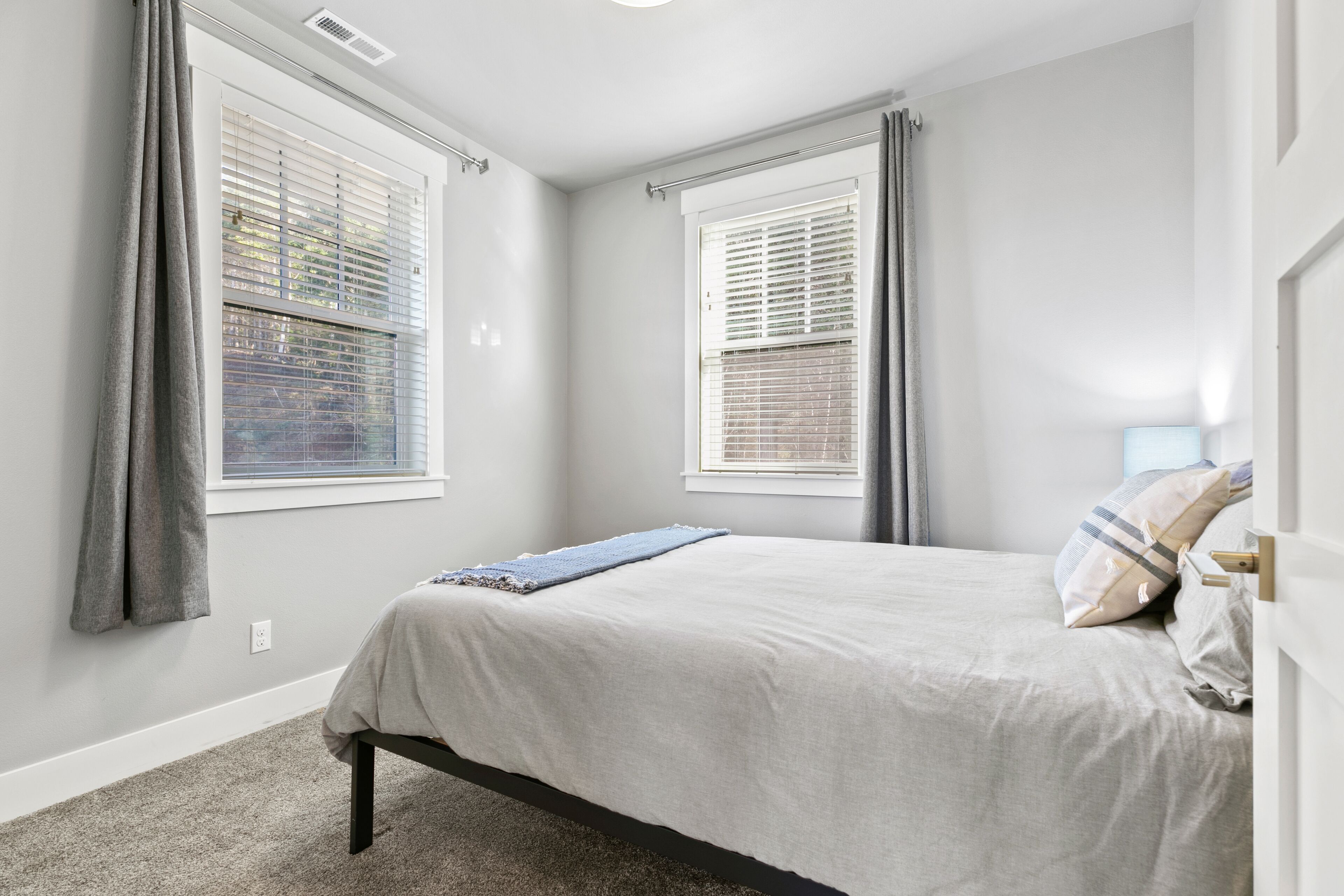 A bedroom with a bed, two windows with blinds, a door, and a carpeted floor.