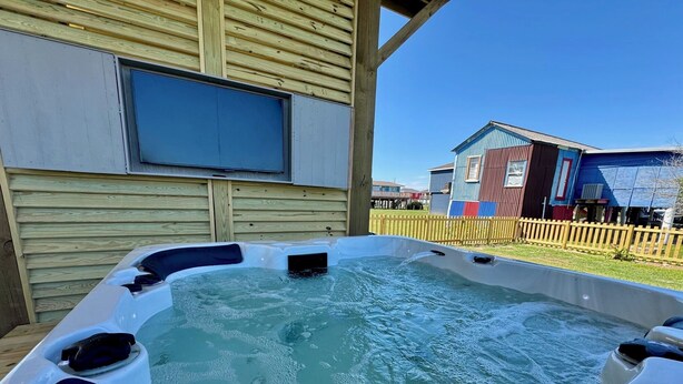 A hot tub with a wooden deck and a building with a large window in the background. - The Sea Glass Retreat - Surfside Beach Texas (Freeport)
