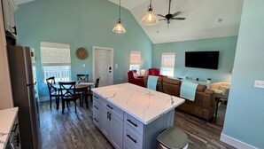 A spacious kitchen with a marble island, a dining area with wooden chairs, a ceiling fan, and a TV mounted on the wall. - The Sea Glass Retreat - Surfside Beach Texas (Freeport)