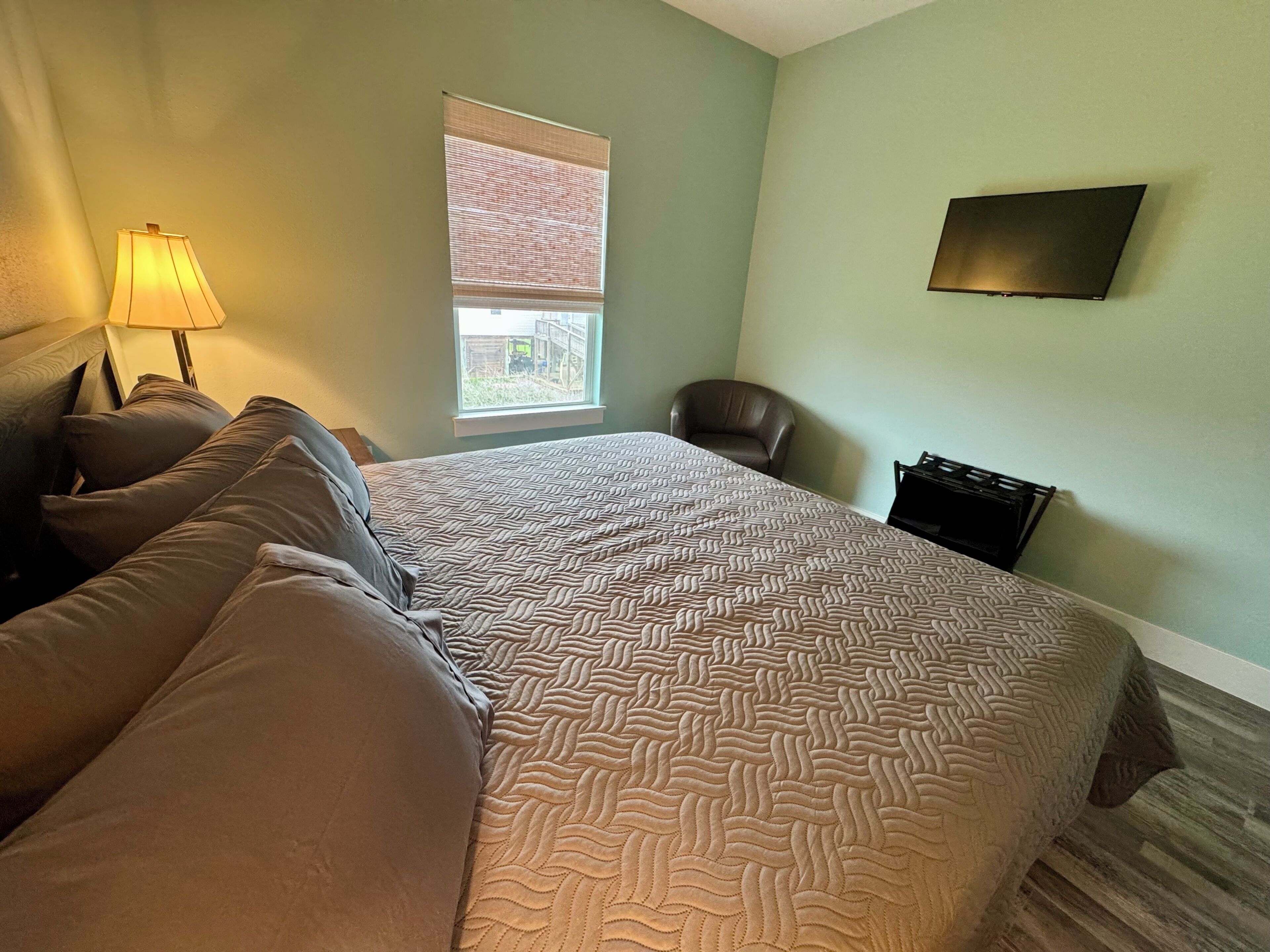 A bedroom with a bed, pillows, a lamp, a chair, a window with blinds, and a TV mounted on the wall.