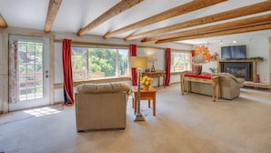 Ein geräumiges Wohnzimmer mit einer Couch, einem Tisch mit Lampe und einem Fernseher. Große Fenster mit Vorhängen, ein Kamin und freiliegende Holzbalken an der Decke.