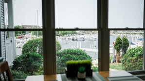 A view from a window overlooking a marina with boats docked, greenery, and a building in the background. - First Floor Condo at Water's Edge (Manistee)