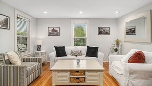 A living room with a white sofa, a striped sofa, and a white armchair. There are two windows with blinds, a lamp, and framed pictures on the walls. - Family Home Near Beach, Downtown, Marina (Manistee)