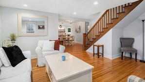 A spacious living room with a wooden staircase, white furniture, and a view into the dining area. - Family Home Near Beach, Downtown, Marina (Manistee)