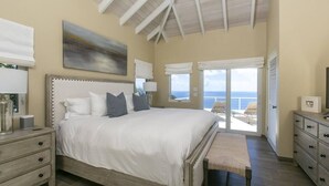 Un dormitorio con una cama grande, un banco, un aparador y vista al océano a través de una puerta corrediza.