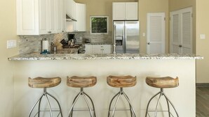 Una cocina con armarios blancos, un refrigerador de acero inoxidable y una encimera de mármol. Hay tres taburetes de madera con patas de metal.