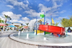 Un parque acuático con un tobogán de agua rojo, múltiples fuentes y un área de piscina con sillas de descanso y sombrillas.