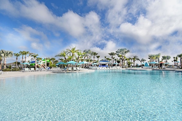 A large outdoor swimming pool with clear blue water, surrounded by palm trees and lounge chairs under blue umbrellas.