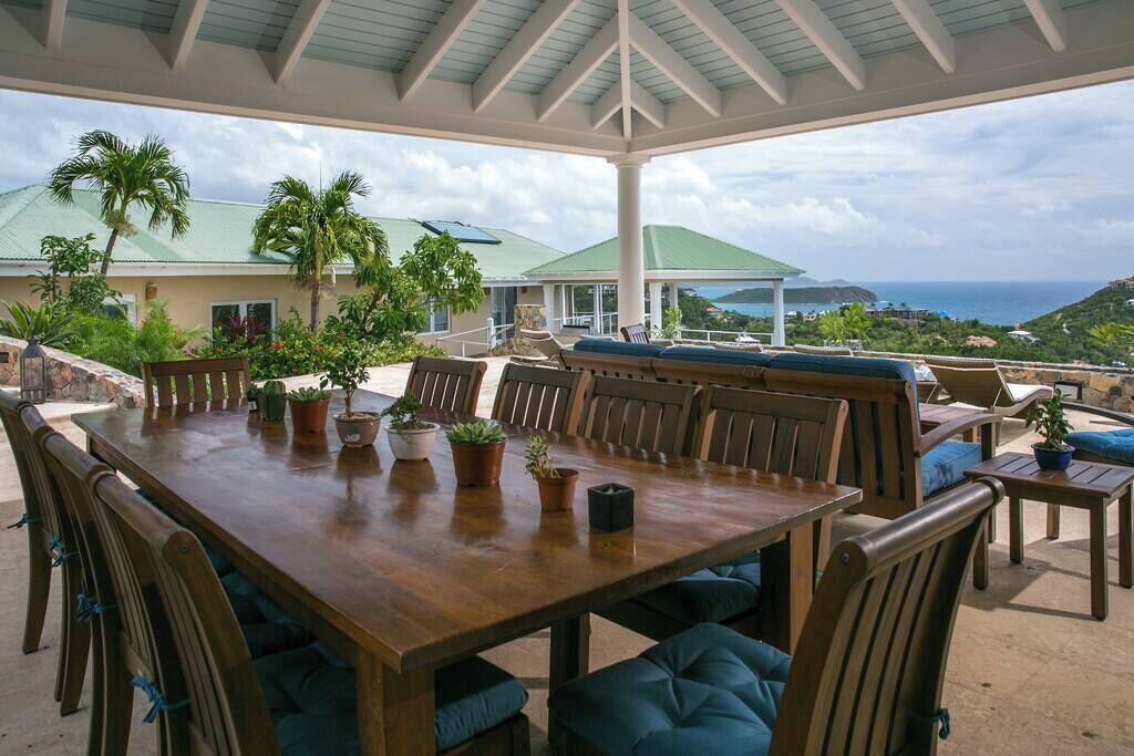 An outdoor dining area with a wooden table and chairs, surrounded by potted plants and a view of the ocean.
