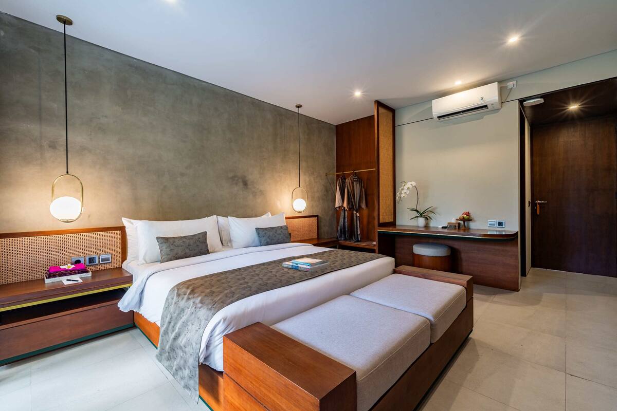 A modern hotel room with a large bed, bedside tables, a vanity with a mirror, and an air conditioning unit.