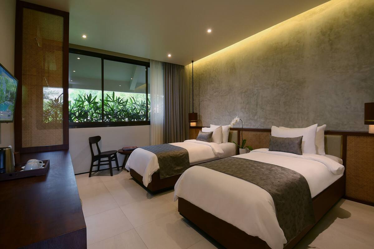 A modern hotel room with two beds, a desk, and a view of greenery outside.
