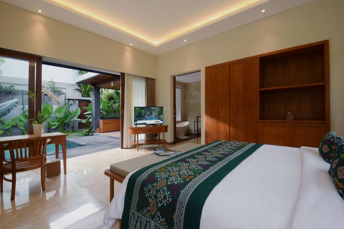 A modern bedroom with a large bed, a desk with a TV, and a sliding door leading to a balcony with plants.