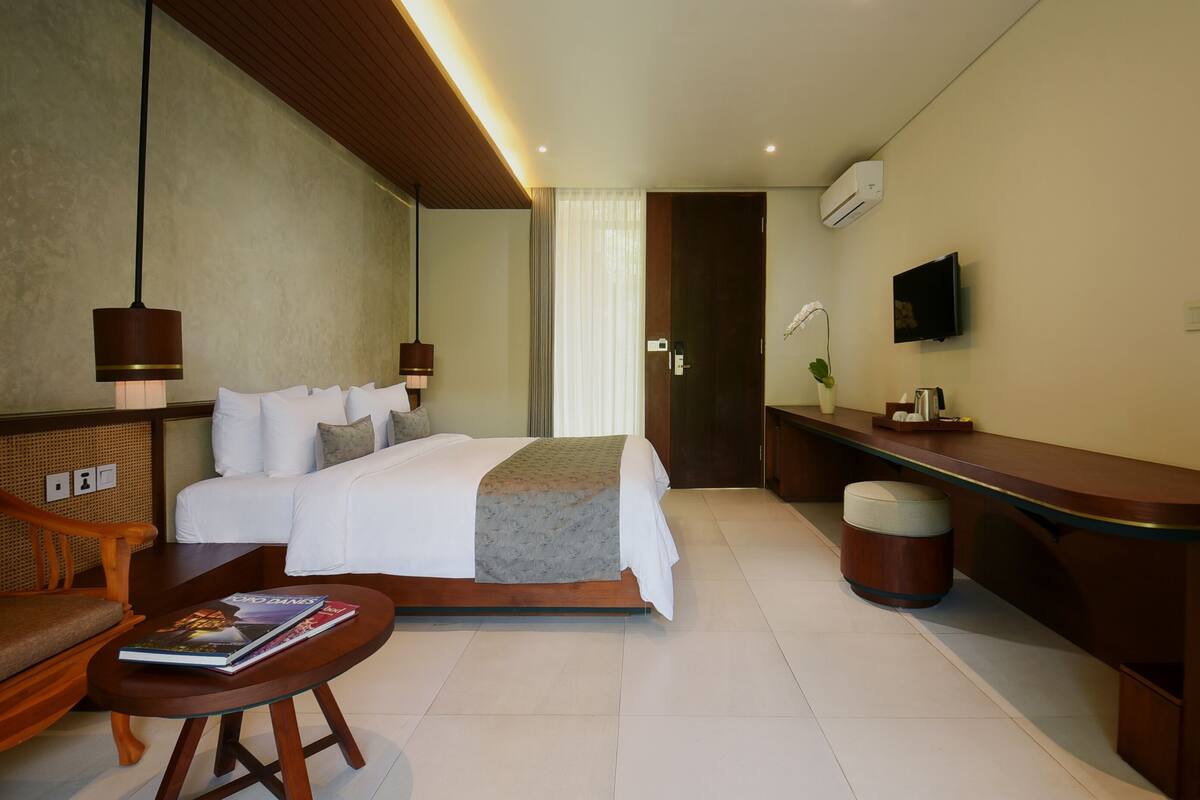 A modern hotel room with a neatly made bed, a desk with a chair, a small table with books, and a TV on the wall.