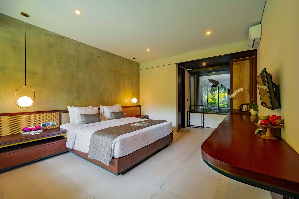 A modern hotel room with a large bed, bedside tables, a wooden desk, and a view of greenery outside.