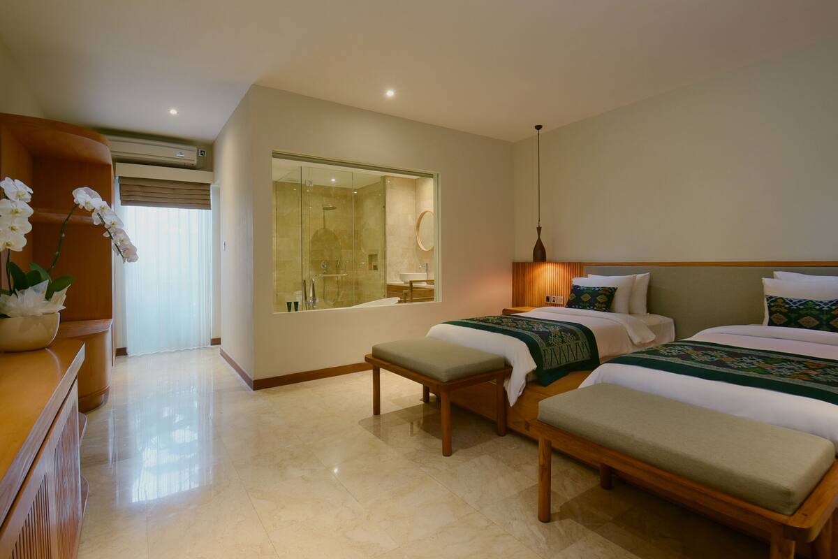 A hotel room with two beds, a sitting area, a large mirror, and a bathroom visible through a glass partition.