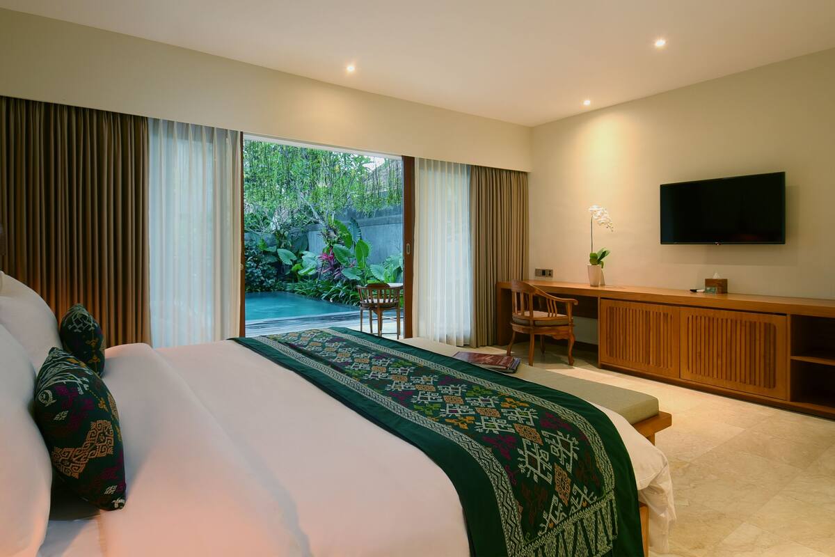A hotel room with a large bed, a desk, and a TV. There is a sliding door leading to a garden.