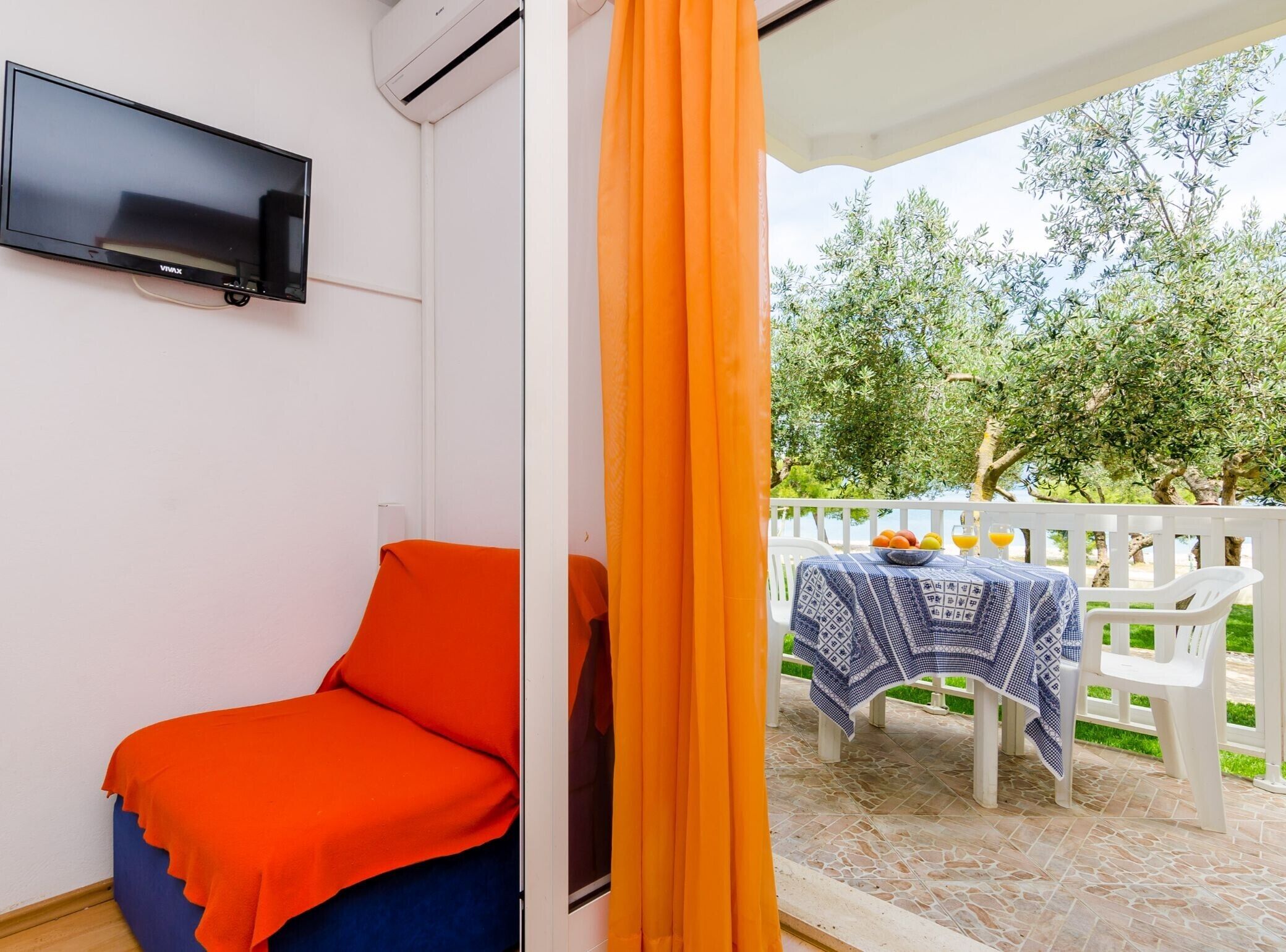 A room with a TV mounted on the wall, a bed with an orange blanket, and a view of an outdoor area with a table and chairs.