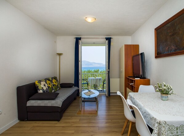 Een knus kamertje met een bank, een eettafel en een televisie. Een balkon met een tafel en stoelen is zichtbaar, met uitzicht op zee.