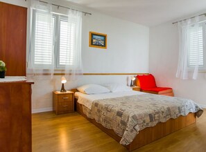 Een slaapkamer met een houten bed, een nachtkastje, een bedlampje, een rode stoel en een raam met witte gordijnen.