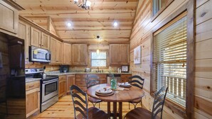 Cocina de cabaña de madera con una mesa redonda, sillas, un refrigerador, estufa, microondas y una ventana con persianas.
