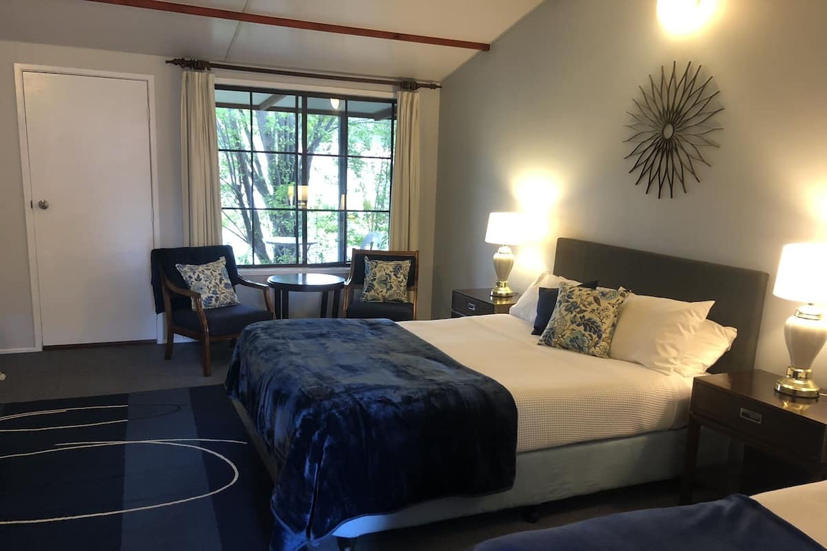 A hotel room with a large bed, two bedside tables with lamps, a small round table with two chairs, and a view of greenery through a window.