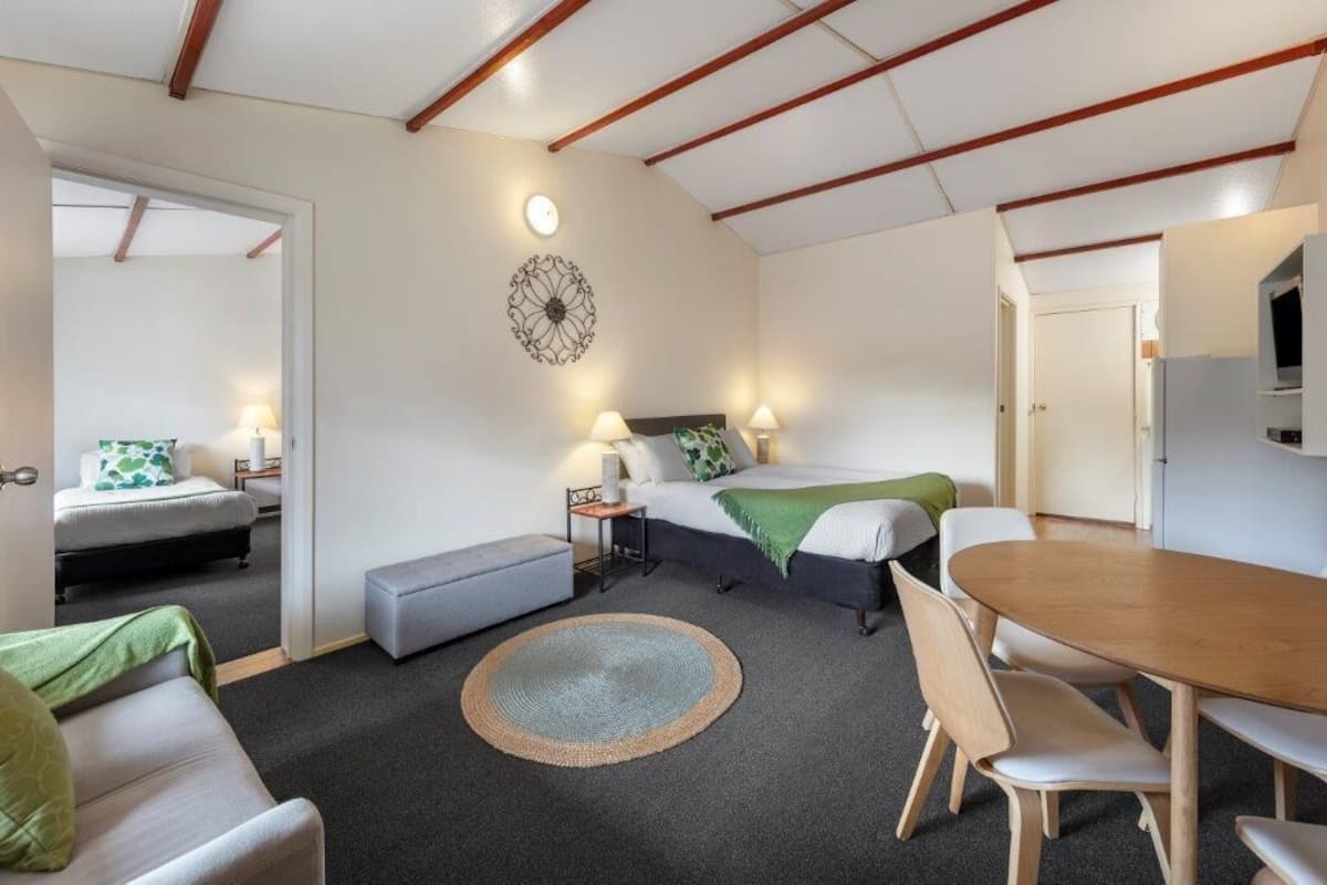 A room with a bed, bedside table, lamp, and a dining table with chairs.