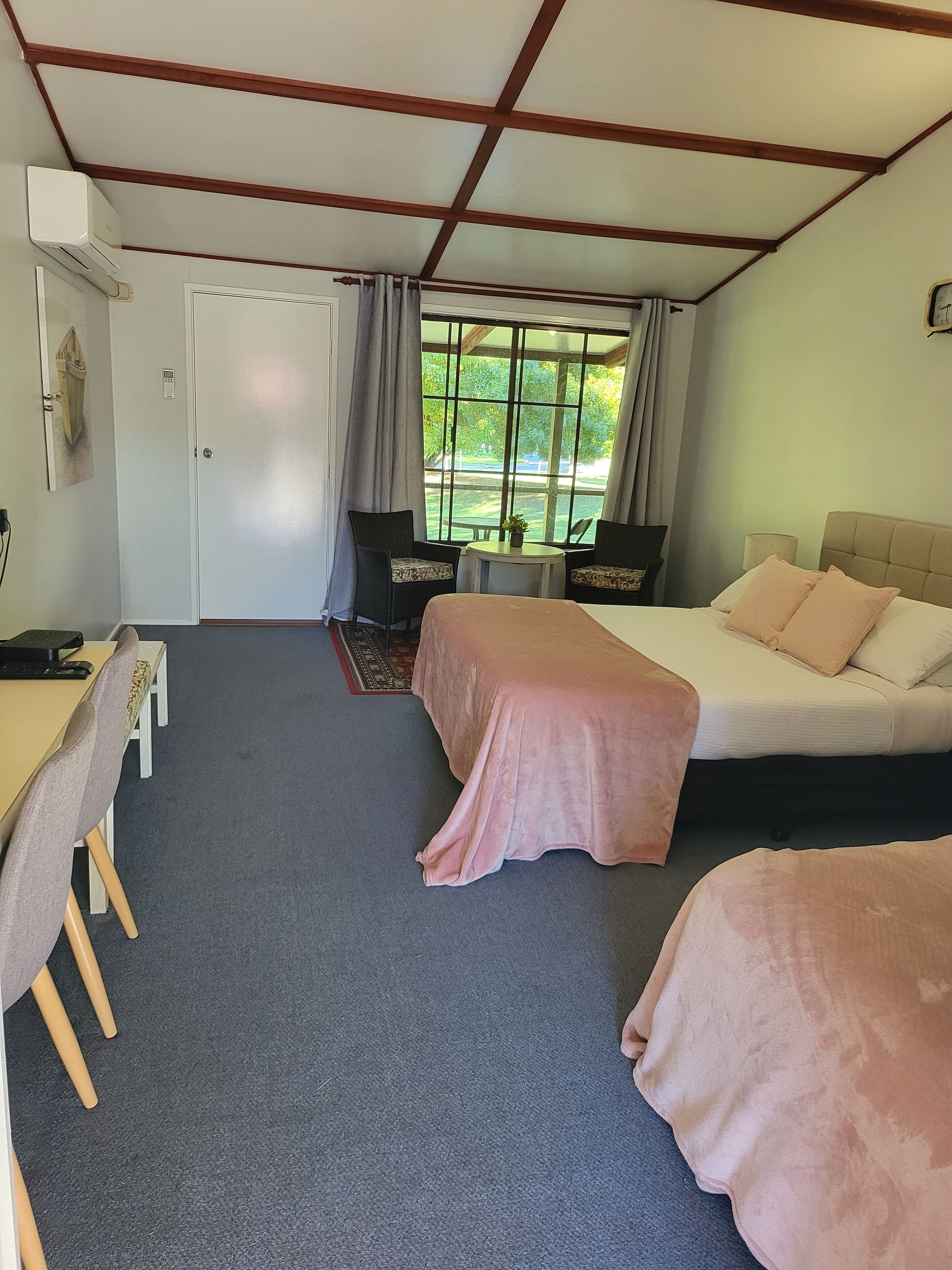 Hotelkamer met een bed, een bureau en een stoel. Er is een raam met uitzicht op bomen en een deur met lichtschakelaar.