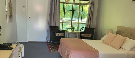 Hotelkamer met een bed, een bureau en een stoel. Er is een raam met uitzicht op bomen en een deur met lichtschakelaar.