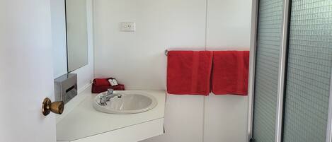 Un baño con lavamanos blanco, espejo y toallas rojas. Hay un cubo de basura y una puerta con mosquitero.