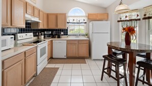 A modern kitchen with wooden cabinets, a white refrigerator, a coffee maker, and a dining area with a round table and bar stools. - PROMO 5BR Villa with Pool/Spa, Near Disney - by Blue Sky Villas (Kissimmee)