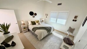 Une chambre à coucher comprenant un lit, des tables de chevet, une commode, une plante en pot, une chaise, une fenêtre avec des stores et un ventilateur de plafond.