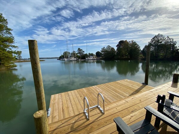 Un muelle de madera con una escalera metálica y bancos, con vista a un agua tranquila con veleros y árboles.