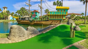Um edifício colorido em formato de barco, com fachada verde e amarela, localizado à beira de um corpo d'água com palmeiras e céu claro.