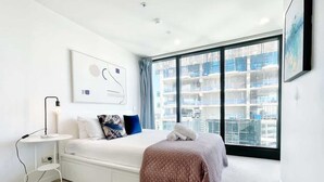 Un hotel moderno con una cama grande, una mesita de noche, una obra de arte colgada en la pared y vistas de un paisaje urbano a través de un ventanal amplio.