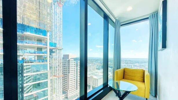 Habitación en un rascacielos con sofá amarillo, una mesa de vidrio, ventanas amplias con vista a la ciudad y una grúa en el exterior.