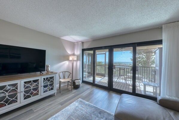 A spacious living room with a large flat-screen TV, a cushioned sofa, and a balcony with outdoor furniture.