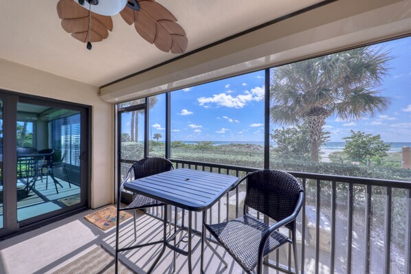 Un balcon avec une table et des chaises, de grandes fenêtres donnant sur des palmiers et l’océan, et un ventilateur de plafond.