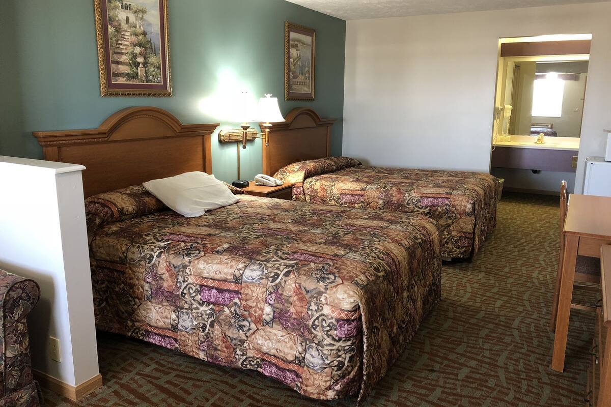Camera d'albergo con due letti, un comodino con lampada, una testiera in legno e un piccolo tavolo.