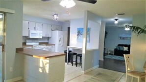 A modern kitchen with white cabinets, a microwave, and a refrigerator. There is a small island with stools and a dining area with a chair and a table.
