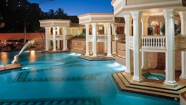 Una piscina con varias construcciones, incluyendo un pabellón y una zona de hidromasaje, rodeada de un jardín ajardinado.