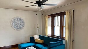 Een kamer met een blauwe bank, een klein witte tafel, een plafondventilator, een decoratief bord aan de muur en een raam met gordijnen.