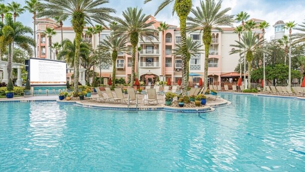 Zona de piscina con sillones, palmeras y un edificio con balcones.
